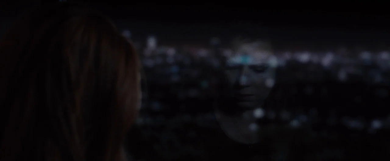 Nocturnal Animals (2016) movie still: closeup — A dark room overlooks a blurred city grid at night through a glass window. A woman stands in the…