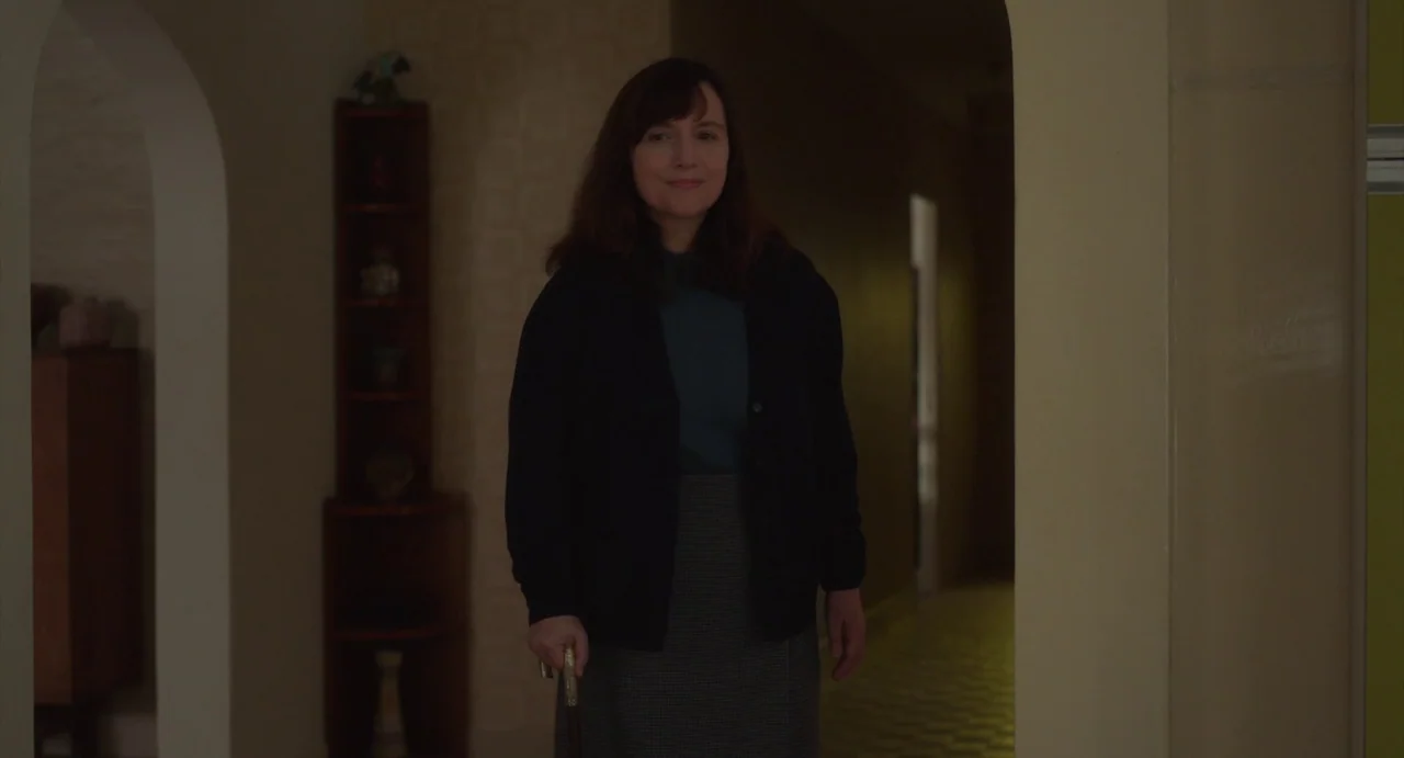 Petite Maman (2021) movie still: medium — A residential hallway with arched doorways and tan walls. A middle-aged woman stands centered in the…