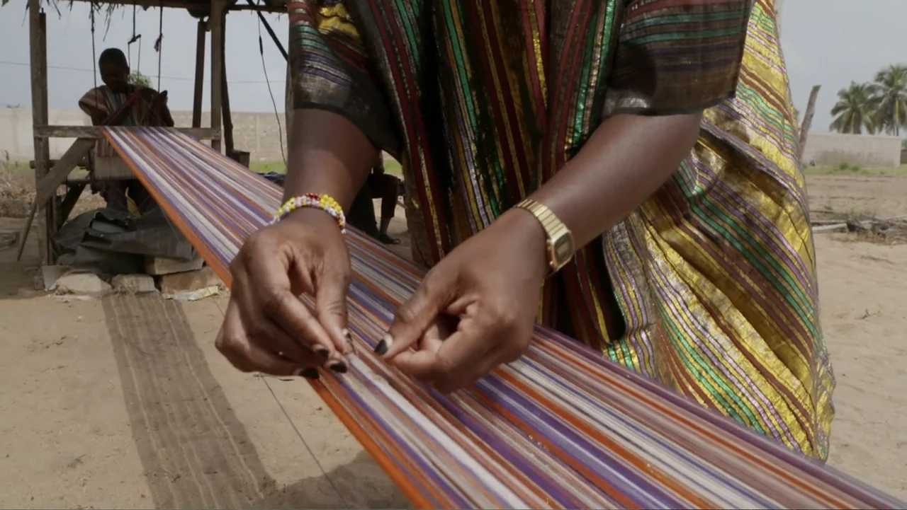 Victoria's Secret: The Tour '23 (2023) movie still: closeup — A wooden loom stands in a sandy exterior area under a simple canopy. A person's hands…