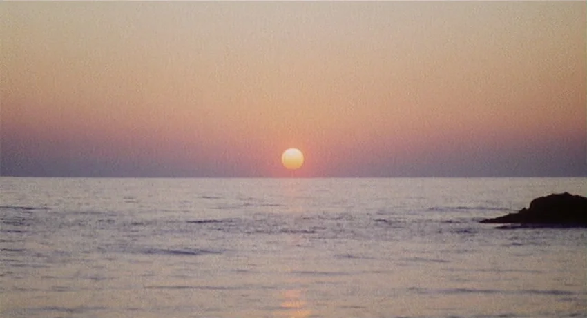 5 Dolls for an August Moon (1970) movie still: establishing — An expanse of calm water reaches toward a clear horizon at dusk. The sun sits…