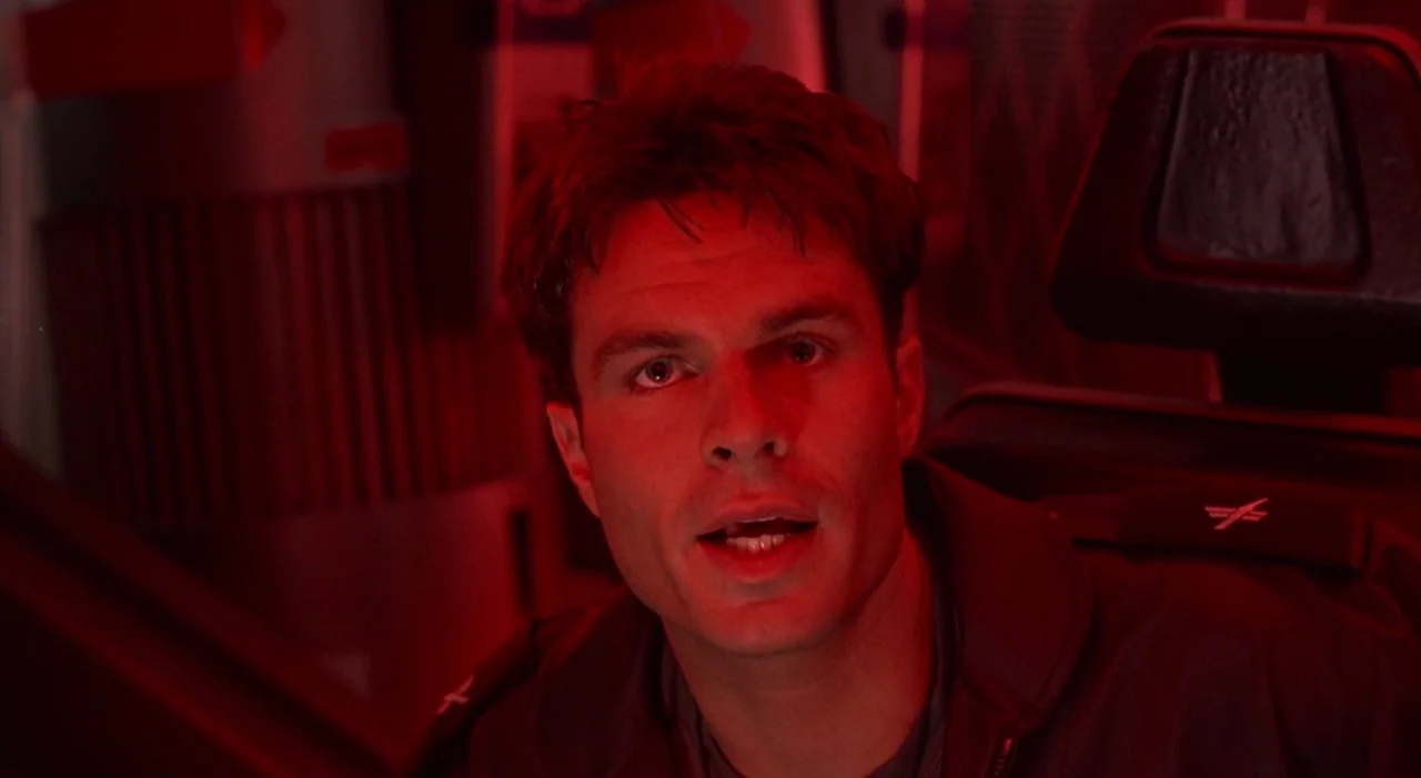 Starship Troopers (1997) movie still: closeup — The cramped interior of a vehicle cockpit is bathed in emergency red light. A young pilot sits in a…
