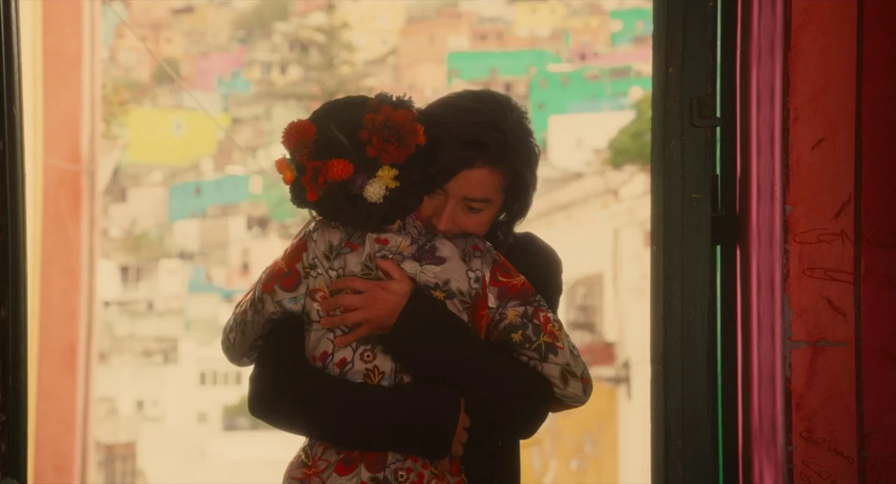 Diner (2019) movie still: medium — A person in a floral shirt and another in black embrace within a doorway. A densely packed hillside neighborhood…