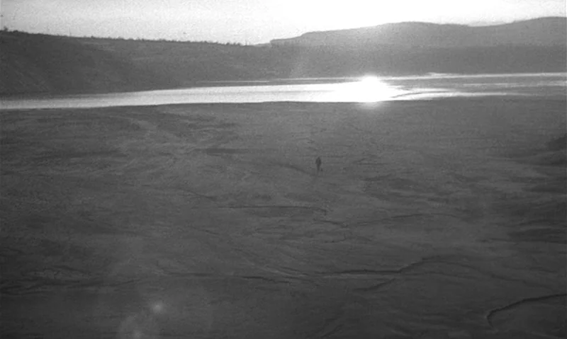 Man Is Not Bird (1965) movie still: wide — A vast, barren mud flat leads toward a reflective body of water under a bright sun. A tiny human figure…