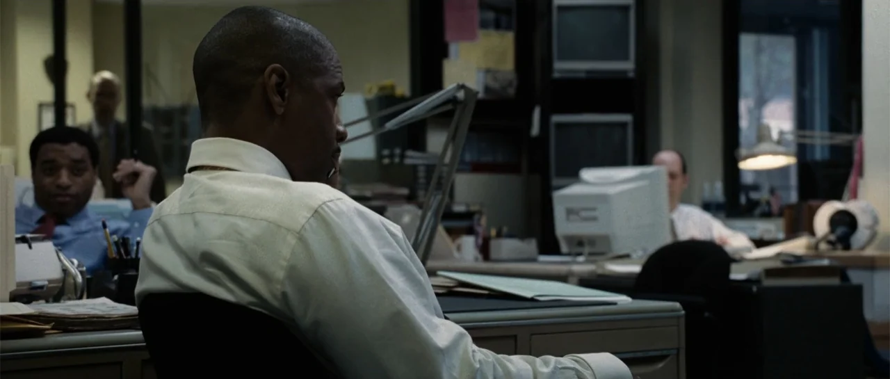 Inside Man (2006) movie still: medium — A cluttered, functional newsroom environment features grey cubicle walls and various desktop electronics. A…