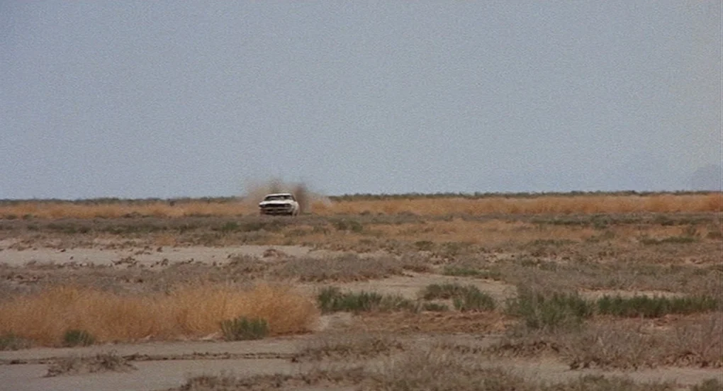 Vanishing Point (1971) movie still: wide — A flat, dry desert plain is covered with patches of withered brown grass. A white car moves across the…