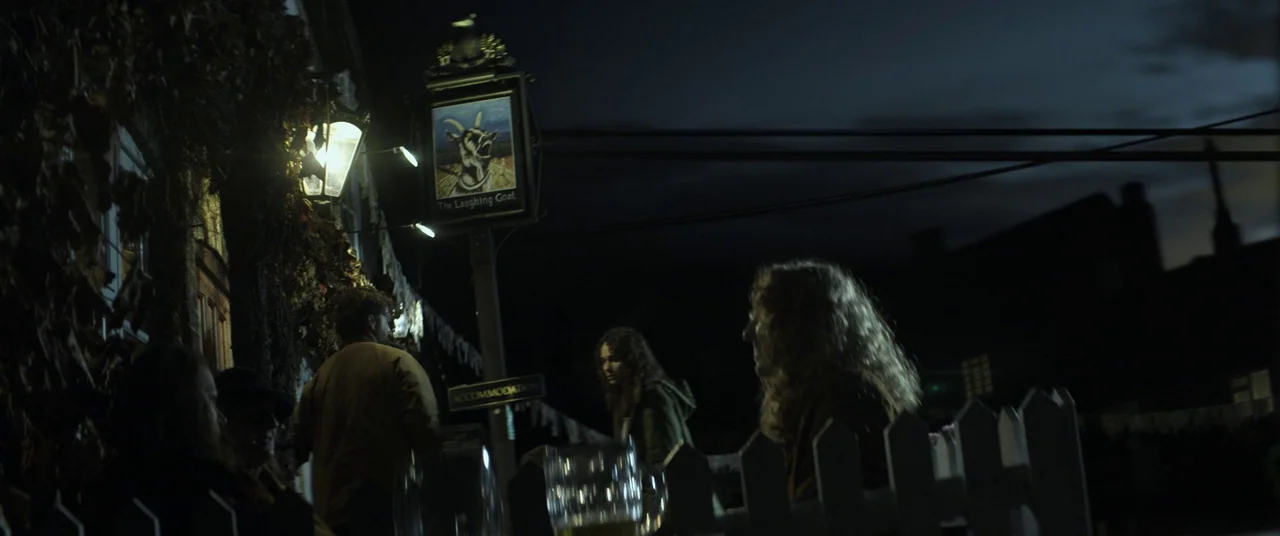 Lord of Misrule (2023) movie still: wide — A pub sign for The Laughing Goat hangs outside a building at night. Several people gather in the shadows…