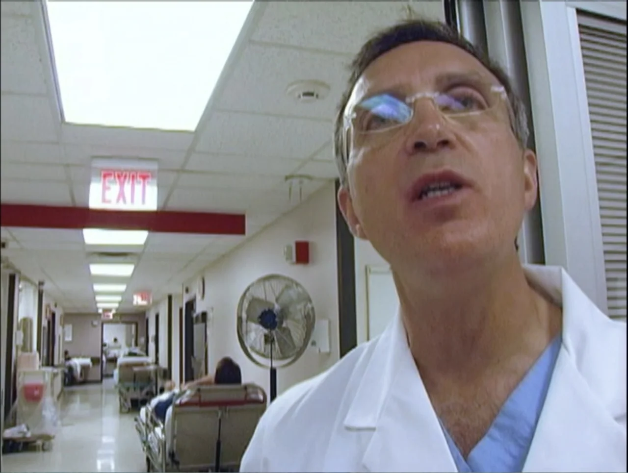 Addiction (2024) movie still: closeup — A long, brightly lit hospital hallway with tiled ceilings and medical equipment visible throughout. A male…