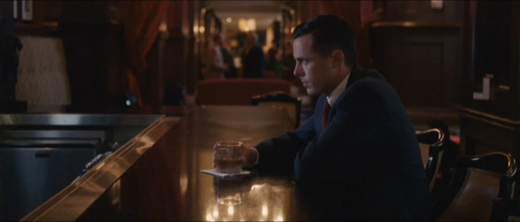 The Killer Inside Me (2010) movie still: medium — A dimly lit, high-end wood-paneled establishment with deep red curtains and blurred figures. A man…