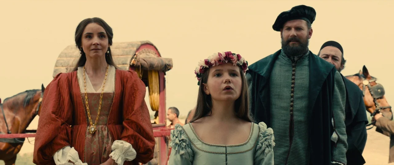 Benedetta (2021) movie still: medium — A group of people in 16th-century style garments stand in an open outdoor setting. A young girl with a floral…