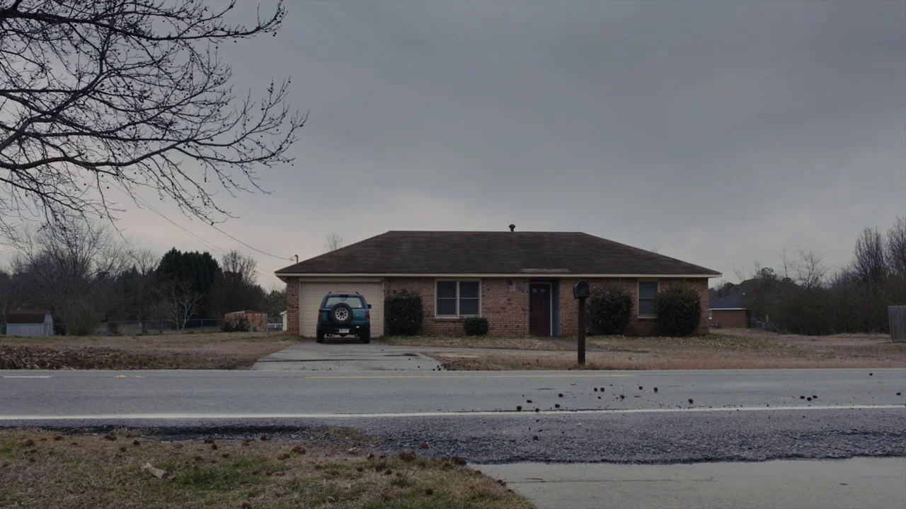 Prisoners (2013) movie still: wide — A single-story brick suburban house sits behind a paved road under a grey sky. An SUV is parked in the driveway…
