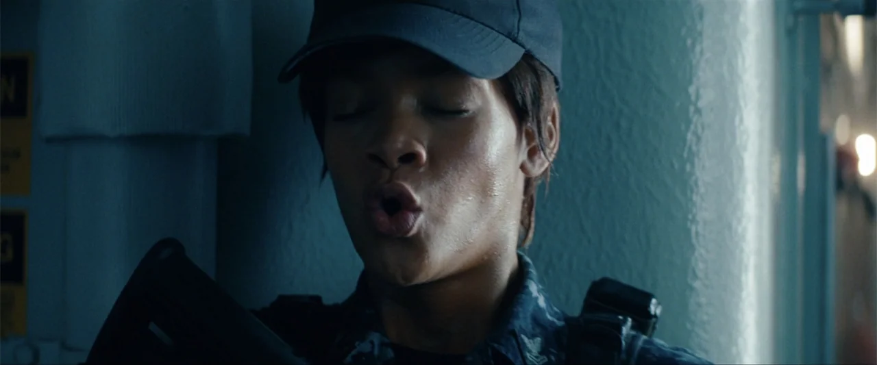 Battleship (2012) movie still: closeup — A dimly lit narrow corridor features blue-toned walls and tactical gear details. A female soldier in…