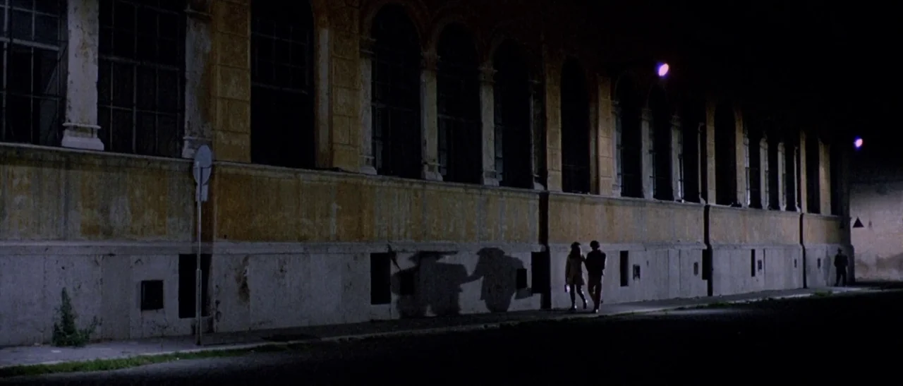 The Bird With The Crystal Plumage (1970) movie still: wide — A long, yellow-painted building with repetitive arched windows sits at night. Two…
