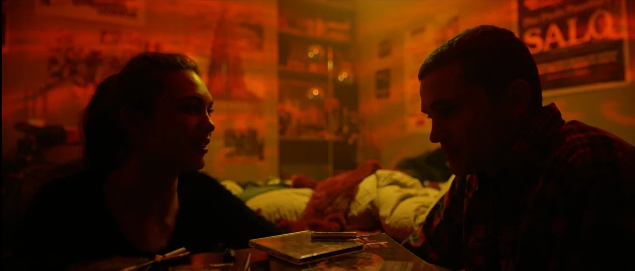 Love (2015) movie still: medium — A dimly lit room is covered in posters and clutter with vibrant red illumination. A man and a woman sit at a low…