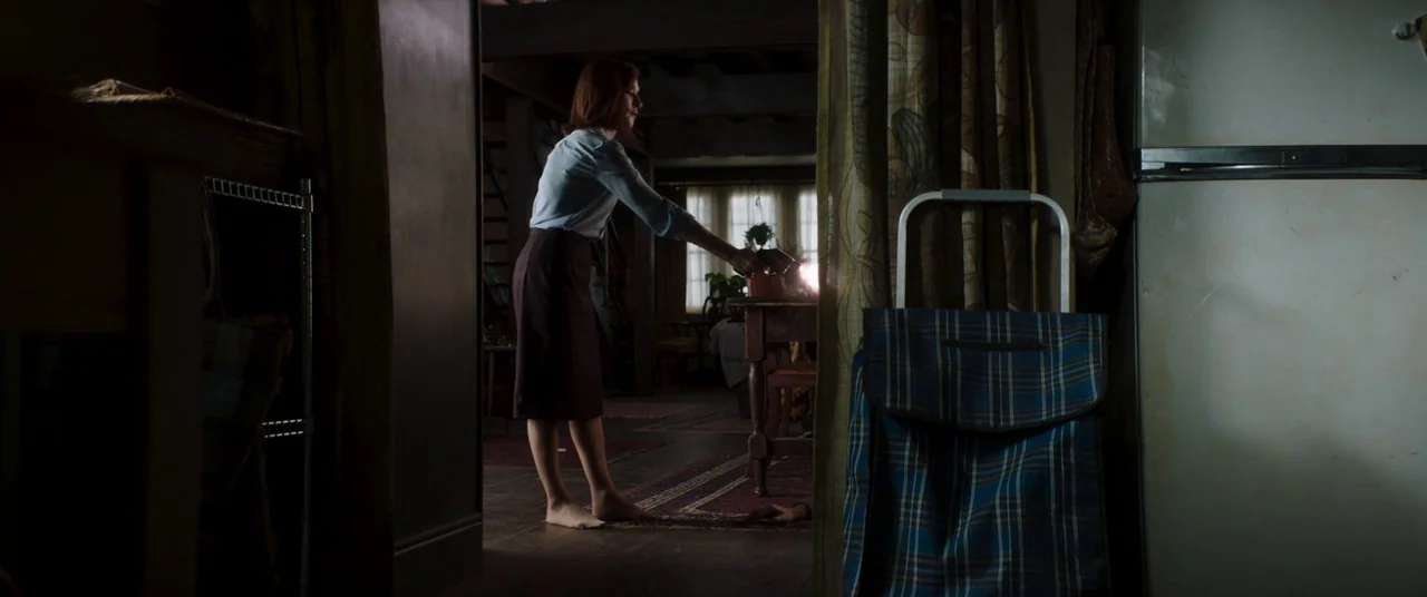 Greta movie still: medium — A dimly lit room with dark wooden floors and vintage patterned curtains near a refrigerator. A woman in a skirt stands…