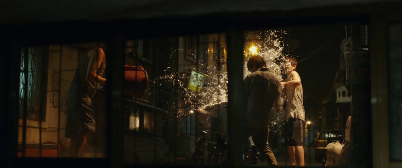 Parasite (2019) movie still: wide — An urban alleyway at night illuminated by distant streetlights. Three young men splash water at each other from…