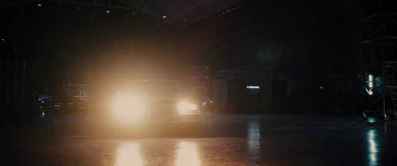 Titane (2021) movie still: wide — A dark, cavernous industrial space contains metal scaffolding and large shipping containers in shadows. A car sits…