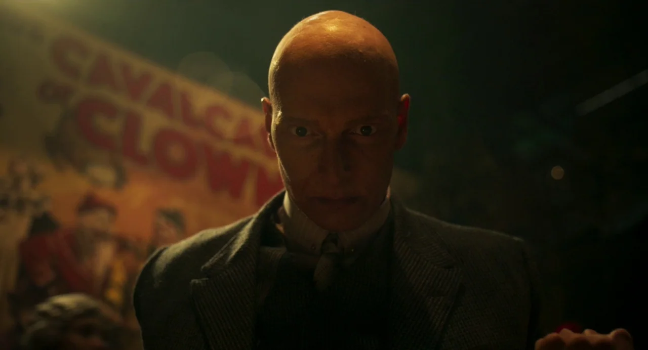 Dumbo (2019) movie still: closeup — A dark, low-lit environment features a vintage poster in the blurred background. A bald man wearing a tweed…