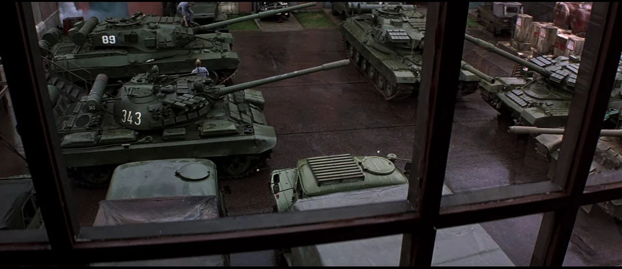 GoldenEye (1995) movie still: wide — A wet concrete lot is filled with rows of green tanks and transport trucks. The view is framed from inside a…