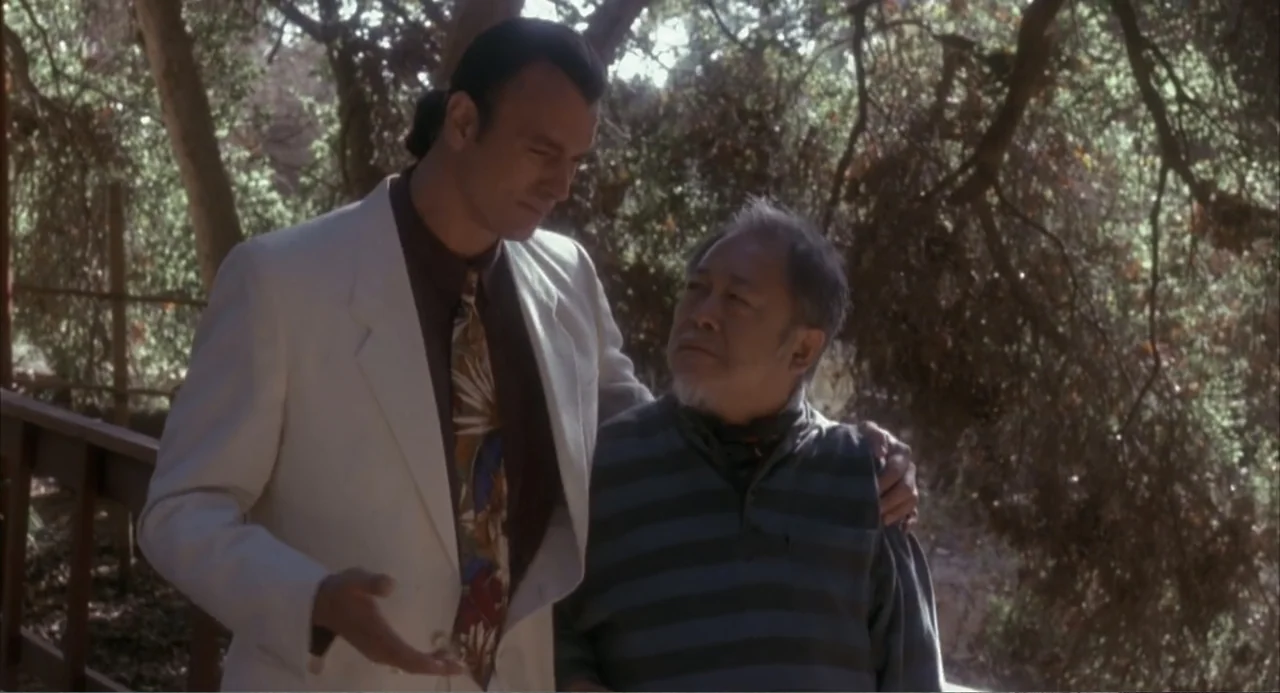 3 Ninjas (1992) movie still: medium — A wooden walkway winds through a grove of trees with dappled sunlight hitting surfaces. A tall man in a white…