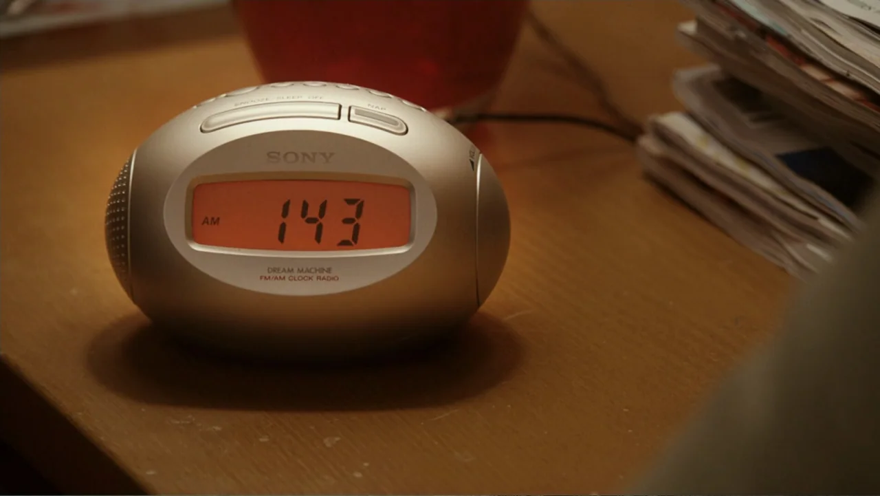 Click (2006) movie still: closeup — A silver Sony Dream Machine clock radio sits on a wooden bedside table. The orange digital display indicates the…