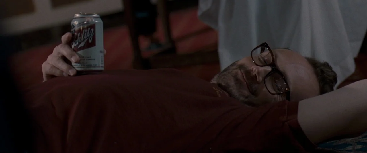 The Innkeepers (2011) movie still: medium — A man in a maroon shirt rests on a floor surface with red carpeting. He holds a can of Schlitz beer…