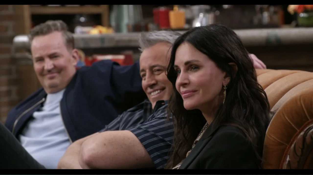 Friends: The Reunion (2021) movie still: medium — A domestic space features a brown upholstered sofa occupied by three adults. The group sits…