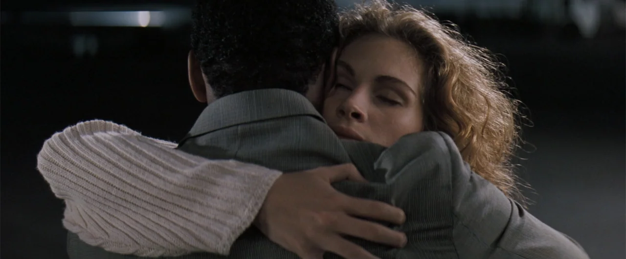 The Pelican Brief (1993) movie still: closeup — A man in a grey suit and a woman in a cream sweater embrace closely. The background is a dark,…