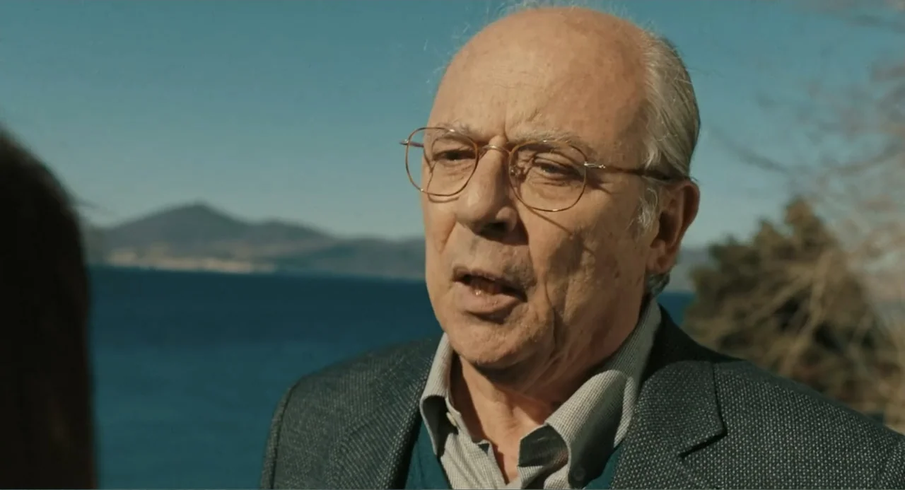 Elles (2018) movie still: closeup — A coastal setting with mountains visible across a body of water in the background. An older man in a blazer…