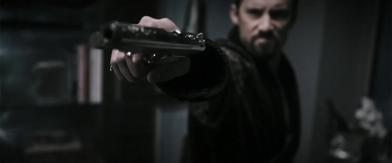 Wild Card (2015) movie still: medium — A dimly lit room contains a man holding a firearm in the foreground. The man stands in an interior space…