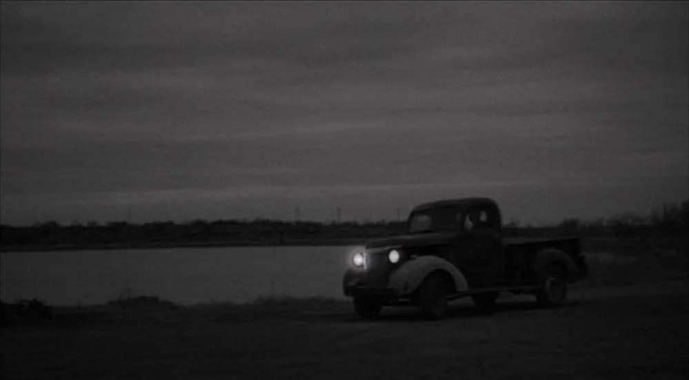 The Last Picture Show (1971) movie still: wide — A rural dirt path runs alongside a dark body of water under a cloudy sky. An vintage 1940s pickup…