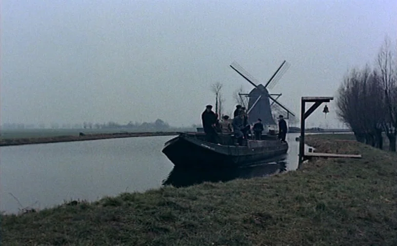 Mill of the Stone Women (1960) movie still: wide — A flat, overcast rural landscape features a narrow canal and a distant windmill. A small wooden…