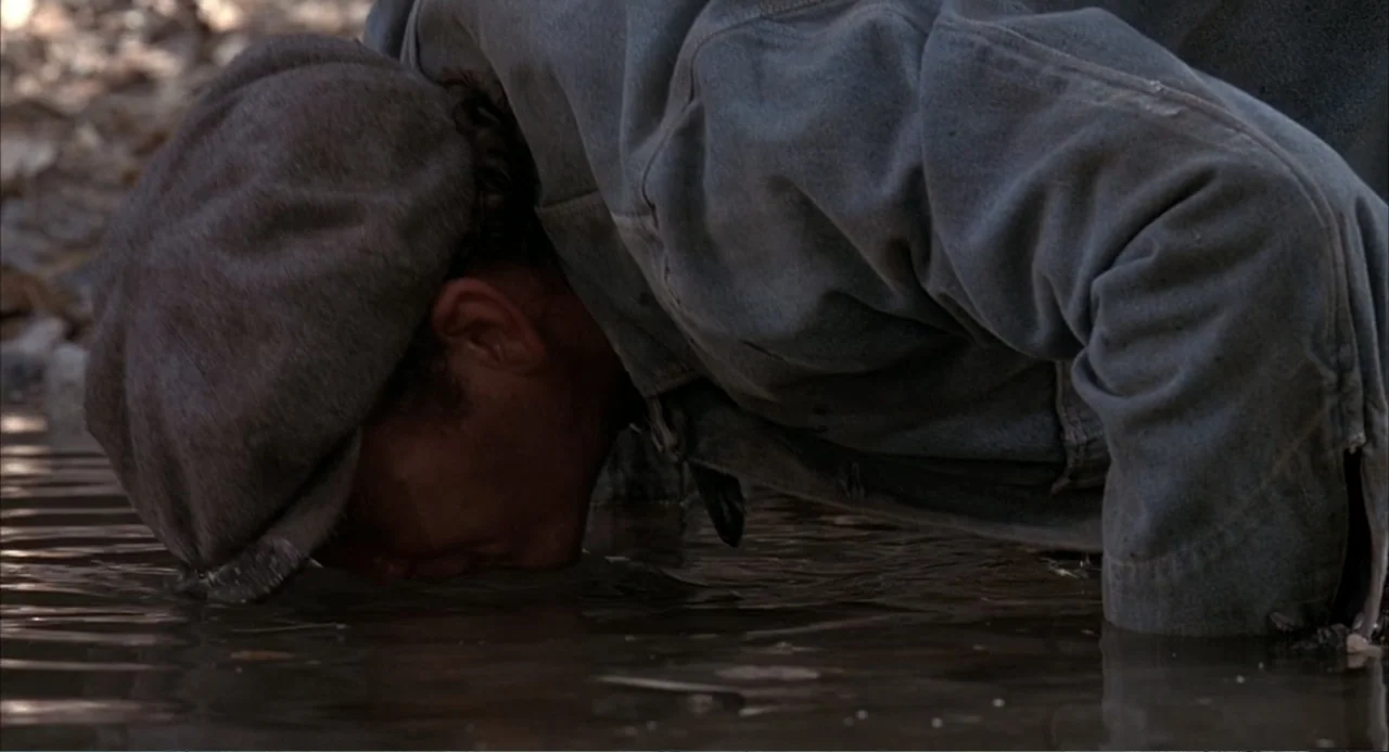 Of Mice and Men (1992) movie still: medium — A man kneels at the edge of a shallow murky stream outdoors. He leans down with his face partially…