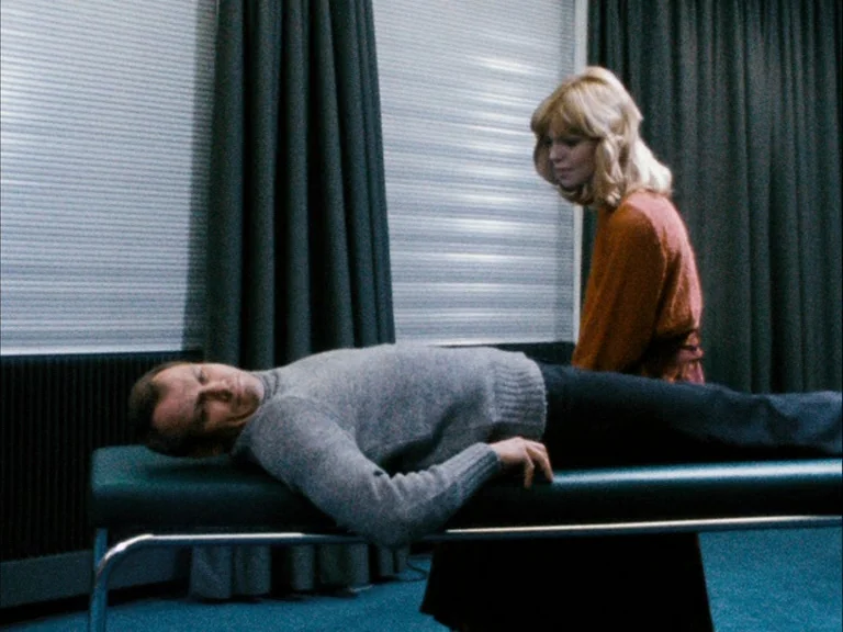 Making of: World On A Wire (1973) movie still: medium — A sparse room features vertical blinds covered by heavy curtains and a low exam table. A man…