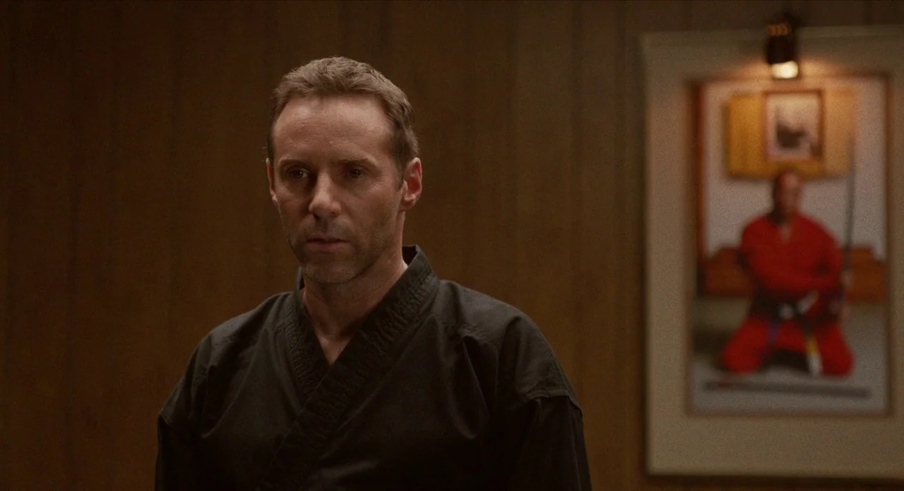 The Art of Self Defence (2019) movie still: medium — An interior space features wood-paneled walls and a framed photo hanging on the wall. A man…
