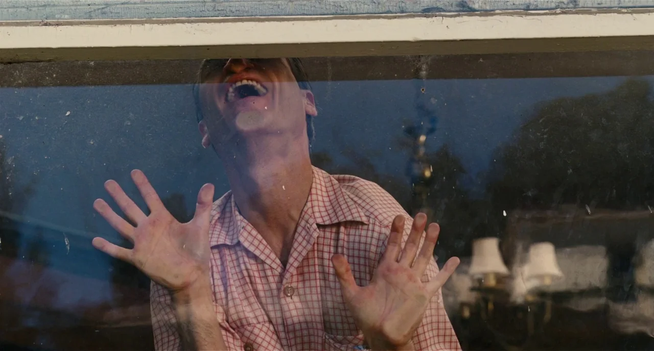 The Master (2012) movie still: medium — A man stands behind a dirty glass window pane, appearing to be outside. He presses his palms against the…