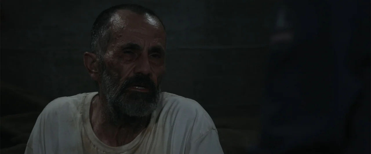 Black Site (2022) movie still: medium — A dimly lit room with dark, featureless walls behind an older man. A man with a disheveled beard wearing a…