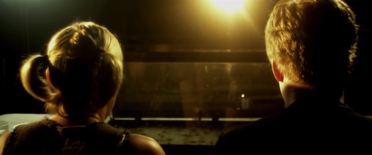 Bellflower (2011) movie still: medium — A dark, dimly lit environment features a glass tank with a warm light source behind. A woman and a man sit…