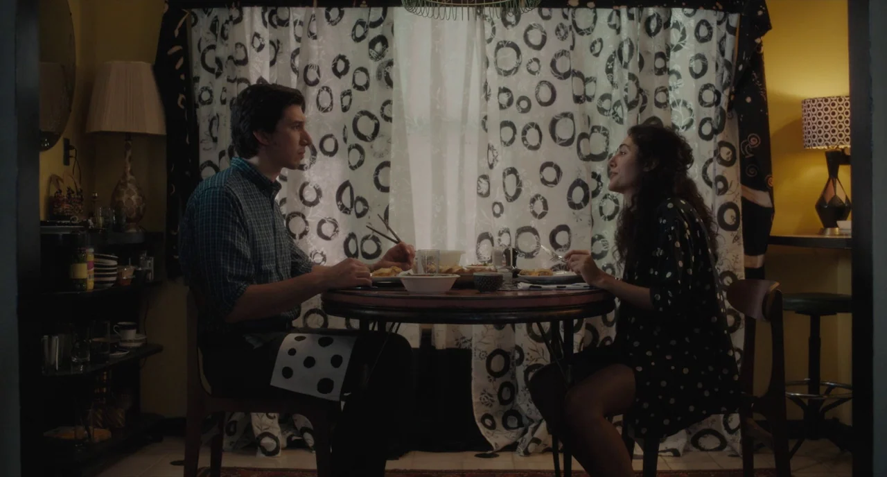 Paterson (2016) movie still: medium — A dining space with yellow walls and prominent black and white circle patterned curtains. A man and a woman…