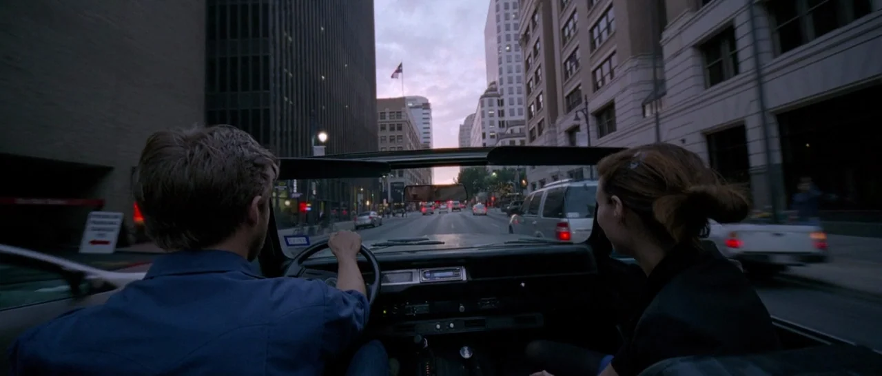 Song to Song (2017) movie still: medium — An urban thoroughfare framed by tall office buildings at dusk with cars in motion. A man drives an…