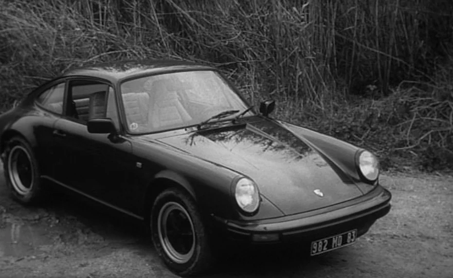 Vivement Dimanche (1983) movie still: fullbody — A classic dark sports car is parked on a rough dirt track by tall reeds. The vehicle is angled…