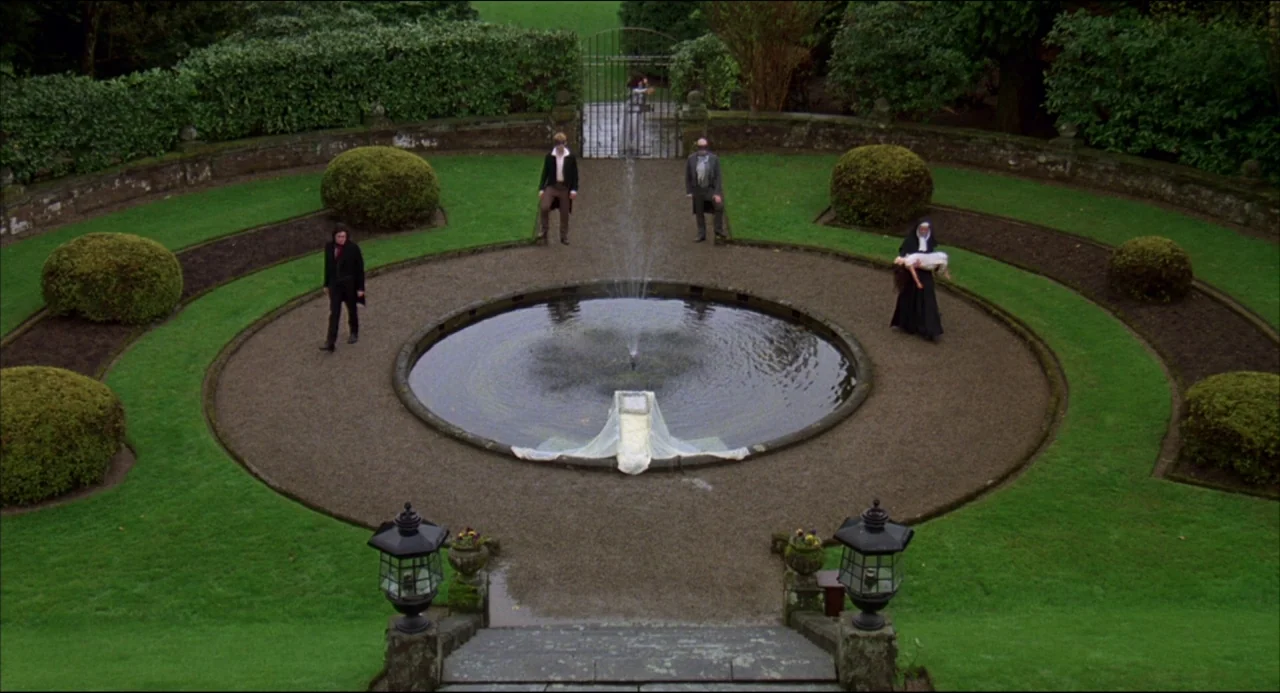 Gothic (1986) movie still: wide — A stone courtyard features a central circular fountain surrounded by manicured hedges and grass. Four individuals…