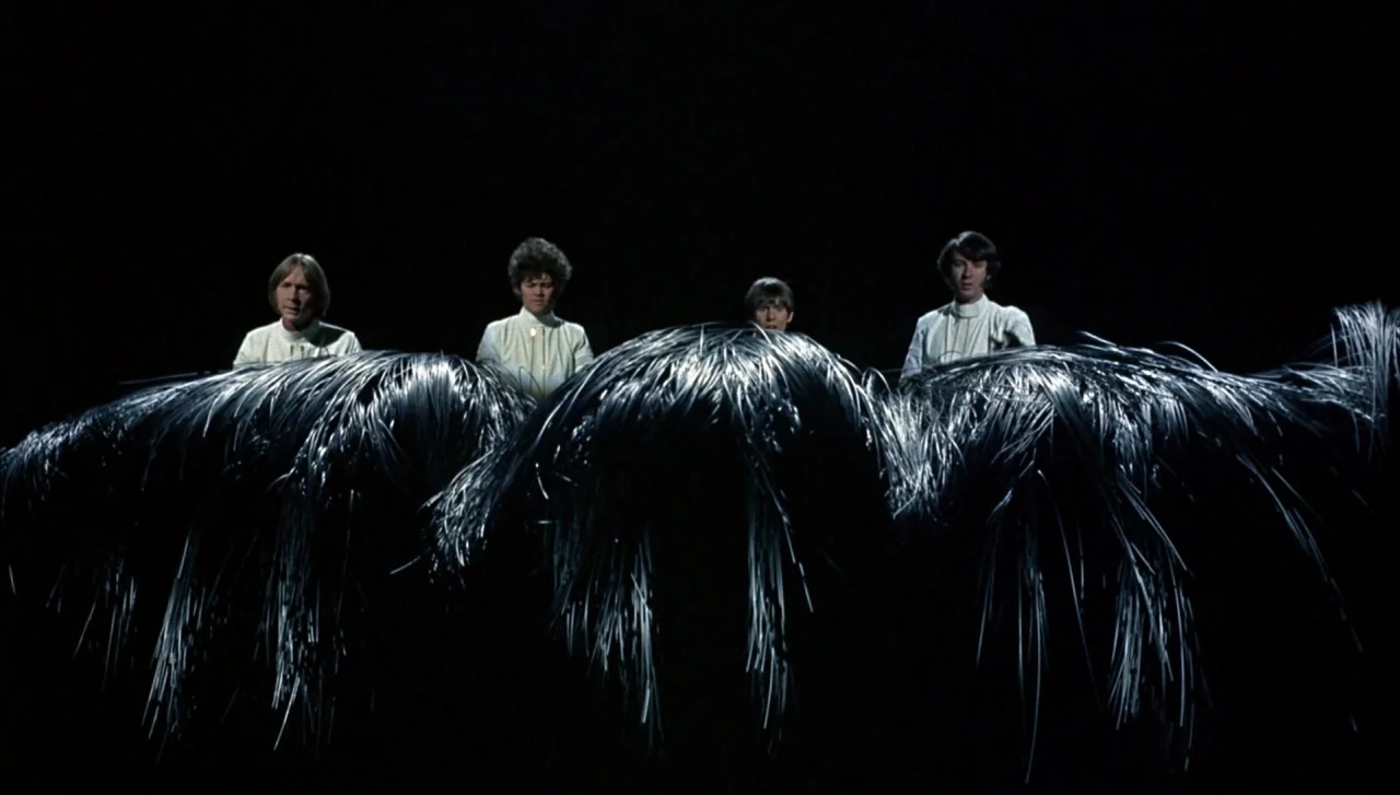 Head (1968) movie still: wide — Four men in white shirts stand behind large, silver, spiky metallic sculptures. The background is a solid black,…