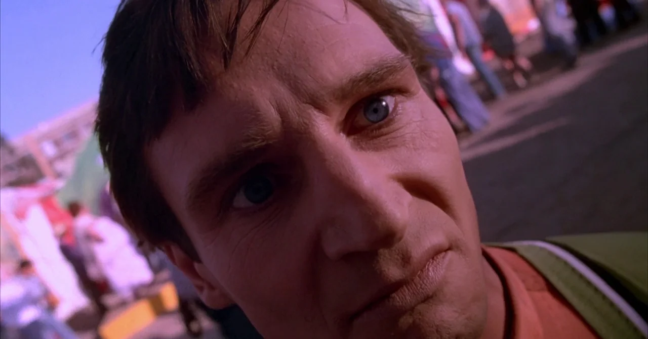 Darkman (1990) movie still: closeup — A man looks down at the camera amidst a blurry, sunlit outdoor event area. He has short hair and blue eyes…