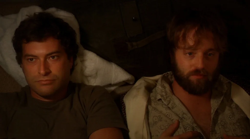 Humpday (2009) movie still: medium — Two men lie next to each other on a dark surface indoors at night. They are both resting in a recumbent…