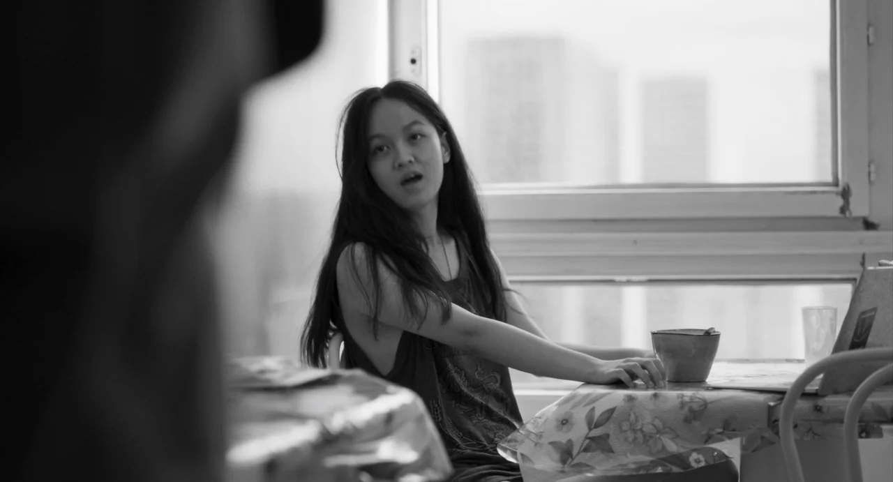 Paris, 13th District (2021) movie still: medium — A sparse interior with a table set against a window overlooking a bright urban exterior. A young…