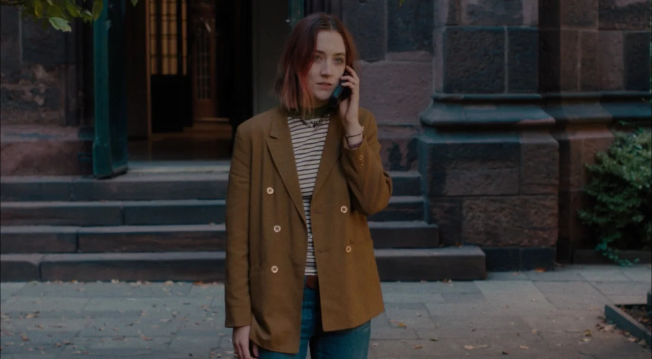 Lady Bird (2017) movie still: medium — A woman stands before the heavy stone steps of a traditional stone building. She holds a smartphone to her…
