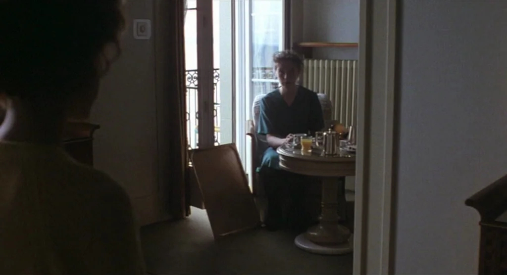 King Lear (1987) movie still: medium — A hotel room with a balcony door and a table set for breakfast. A woman sits in a chair at the small round…