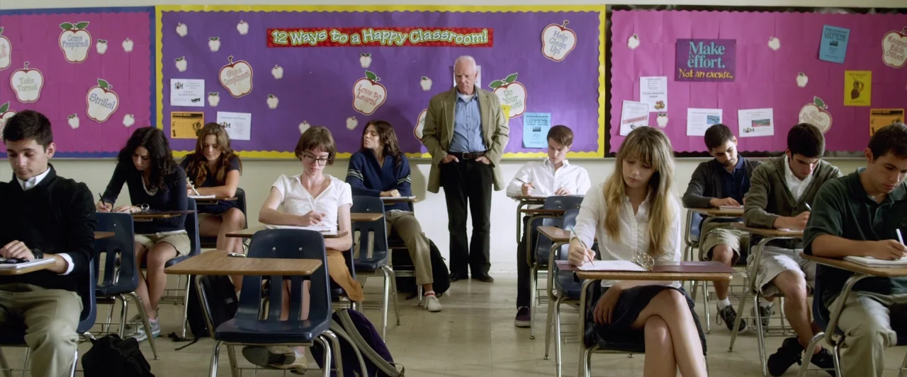 Excision (2012) movie still: wide — A high school classroom features rows of blue plastic desks against purple bulletin board walls. Multiple…