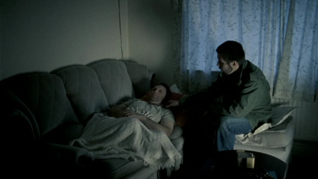 Dead Man’s Shoes (2004) movie still: medium — A residential interior features a grey fabric sofa against a plain wall. A man sits beside another man…