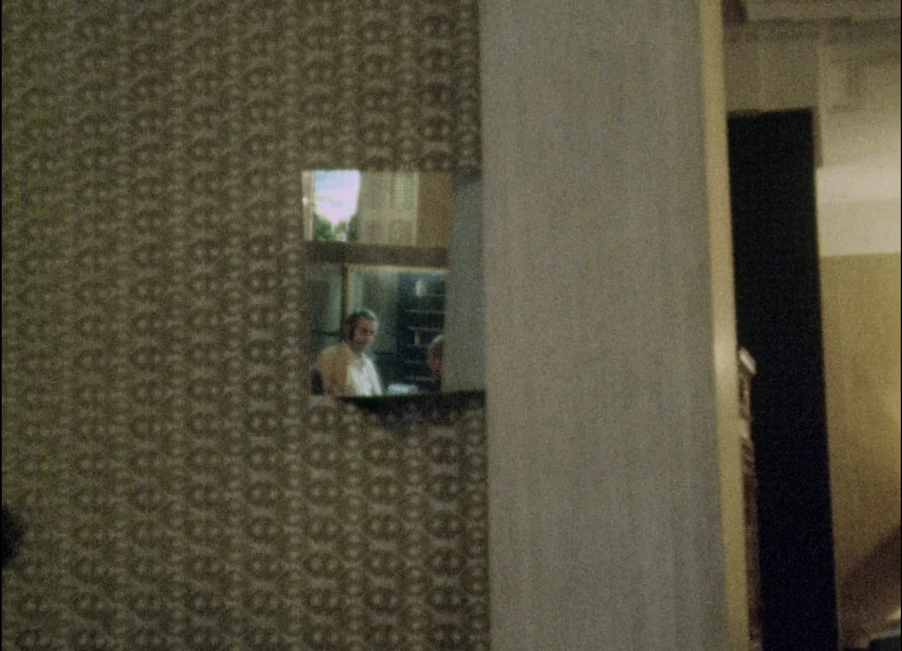 Hotel Monterey (1973) movie still: wide — A hallway with patterned, textured wallpaper leads to an open pass-through window. A man sits at a table…